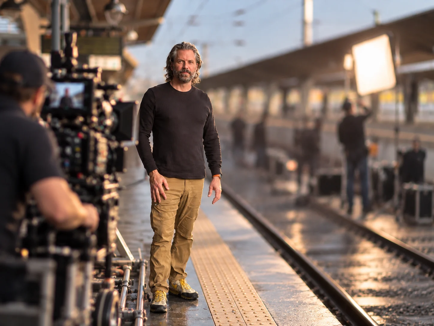 Warren Hansen on set, standing on a rain-wet train platform at golden hour with a cinema camera on a dolly in the foreground and a lighting crew behind him